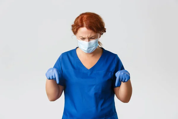 Medical workers, covid-19 pandemic, coronavirus concept. Frustrated frowning middle-aged female doctor, nurse in face mask and gloves frowning confused and pointing fingers down, grey background
