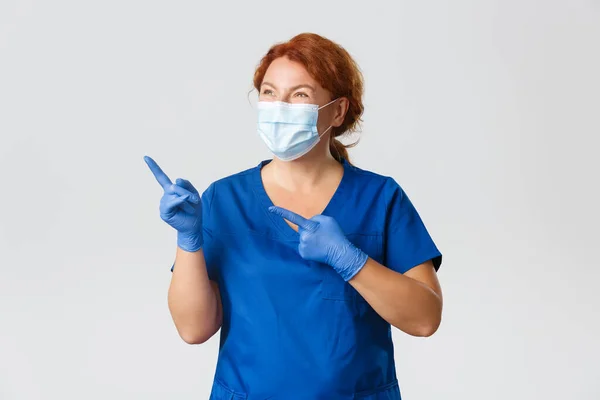 Medical workers, covid-19 pandemic, coronavirus concept. Portrait of happy smiling female doctor in face mask, rubber gloves and scrubs grinning delighted and pointing upper left corner