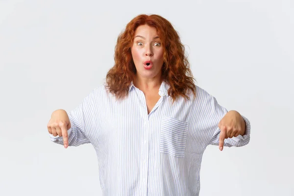 Excited and impressed middle-aged female entreprenuer, redhead woman react to amazing promo offer, pointing fingers down and showing advertisement special price, standing white background — Stock Photo, Image