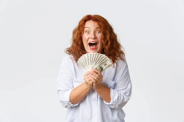 Pengar, finans och människor koncept. Triumferande, framgångsrik och vinnande rödhårig medelålders kvinna jublar över en enorm summa pengar i sina händer, ser glada och glada över vit bakgrund — Stockfoto