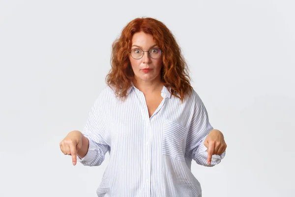 Shocked and speechless middle-aged redhead woman in glasses stare at camera with popped eyes while pointing fingers down at something strange, standing white background amazed — Stock Photo, Image