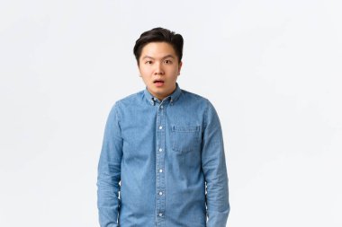 Shocked and startled asian male student, drop jaw and stare dumb at camera, cant understand what happening, feeling confused and puzzled, looking speehless over white background