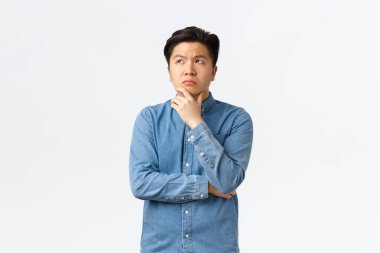 Düşünceli, karmaşık Asyalı bir adam, çenesine dokunuyor ve sol üst köşeye bakıyor, düşünüyor, karar veriyor, bir şeyler seçiyor, şüpheleri var, beyaz arka plan üzerinde tereddütlü duruyor.