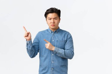 Şüpheci ve öfkeli Asyalı adam sol üst köşeyi işaret ediyor, kötü haberler veriyor, kaşlarını çatıyor, bir şey için azarlıyor, beyaz arka planda durmaktan rahatsız oluyor, ifade veriyor.