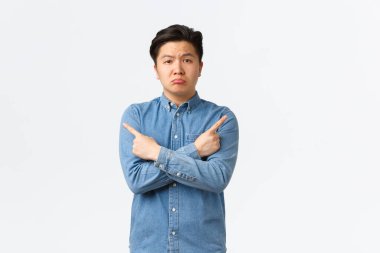 Hüzünlü yakışıklı Asyalı adam somurtuyor, şikayet ediyor, pişmanlıkla yan gözle bakıyor, sol ve sağ varyantları gösteriyor, ikisi de kötü, beyaz arka planda duruyorlar.