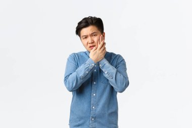 Tedirgin ve rahatsız Asyalı adam acı verici duygulardan yakınıyor, yanağa dokunuyor, diş ağrısı çekiyor, stomatoloji kliniğine gitmesi gerekiyor, randevusu var, ağrı kesicileri bekliyor, beyaz arka planda duruyor.