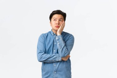 Sıkıcı ve bitkin Asyalı erkek öğrenci kendini eğlendirilmemiş hissediyor, derste boş yer var, yüzünü yana çevirip hayallere dalmış, sıkılmış, beyaz arka planda duruyor, sıkıcı sınıflara katılıyor.