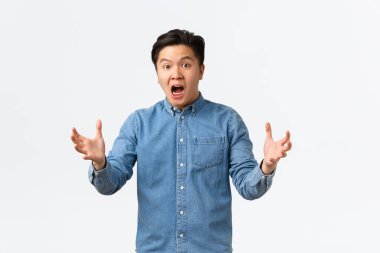 Şok olmuş ve paniğe kapılmış Asyalı bir adam kötü habere tepki veriyor, beyaz arka planda panik içinde duruyor ve kararsız bir şekilde el sıkışıyor, ne yaptığını bilmiyor, kendini işe yaramaz, beyaz arka planda hissediyor.