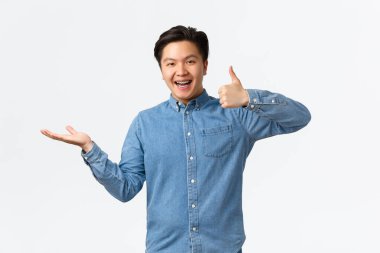 Mavi tişörtlü tatmin olmuş yakışıklı Asyalı adam, sınıflandırma ürünü olarak onayını veriyor, beyaz arkaplanı sanki tutuyormuş gibi kaldırıyor, tavsiye ediyor, bir şeyler gösteriyor.