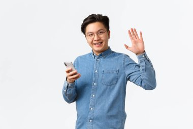 Diş teli takmış gülümseyen Asyalı adam cep telefonu kullanıyor, kameraya bakıyor ve el sallıyor, size merhaba diyor, internetten randevu uygulamasında insanları buluyor, arkadaşlarıyla buluşuyor, beyaz arka planda duruyor.