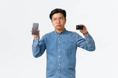 Hayal kırıklığına uğramış ve üzgün Asyalı adam, iç çekerek pişmanlık duyuyor, banka hesabı olmadığından şikayet ediyor, cep telefonu ekranında ve kredi kartında e-bankacılık uygulaması gösteriyor, beyaz arka plan