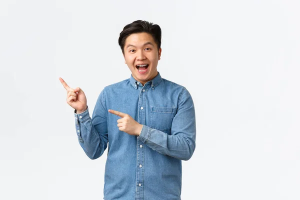 Opgewonden vrolijke Aziatische man in blauw shirt reageren op fantastisch nieuws, wijzende vingers linksboven hoek, demonstreren banner, bevorderen van grote gebeurtenis, glimlachend verbaasd, staande witte achtergrond — Stockfoto