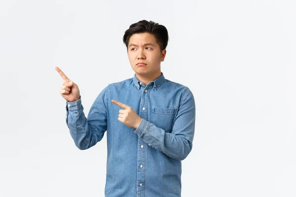 Oroad och besviken asiatisk dyster man i blå skjorta, rynkar pannan upprörd och pekar finger övre vänstra hörnet, klagar dålig produkt, känna sig bekymrad, stående vit bakgrund — Stockfoto