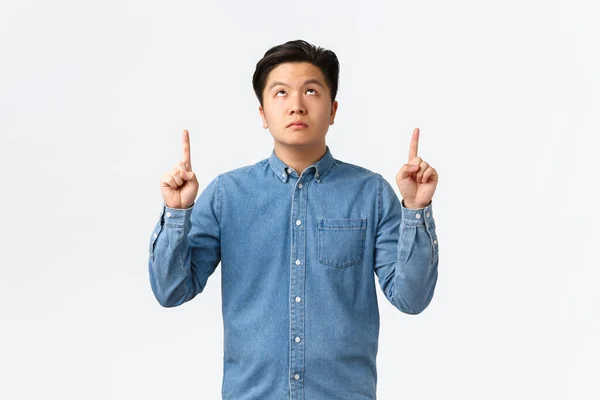 Oroad uttråkad asiatisk kille i blå skjorta, tittar och pekar finger upp med pokeransikte, inga känslor, vara slarvig med information uppåt, läsa tecken utan intresse, vit bakgrund — Stockfoto
