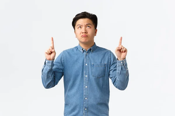 Skeptisk och oroad asiatisk man i blå skjorta tittar och pekar finger upp med besvikna uttryck, sura bedrövad, klagar över något dåligt, stående vit bakgrund — Stockfoto