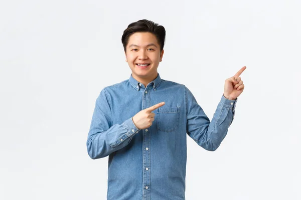 Alegre sorrindo asiático jovem homem com aparelho no azul camisa, apontando dedos superior direito canto orgulhoso e confiante, mostrando link ou fazer anúncio, pé branco fundo — Fotografia de Stock