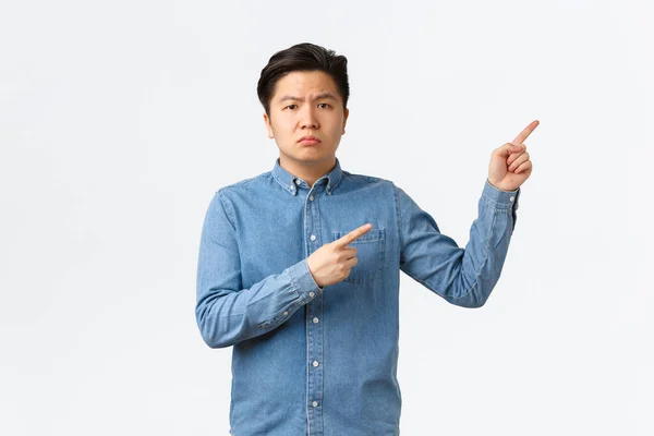 Misstänksam och tveksam asiatisk man i blå skjorta, rynkar pannan skeptisk och missnöjd, pekar finger övre högra hörnet, frågar fråga, klagar på något störande, vit bakgrund — Stockfoto