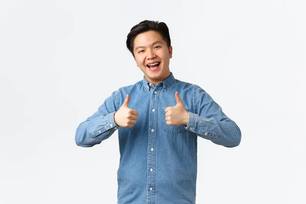 Spokojen šťastný asijský muž s rovnátka, ukazující palce nahoru ve schválení, jako a doporučit produkt nebo službu, gratulace přítel s úspěchem, říká dobře udělal, vynikající, bílé pozadí — Stock fotografie