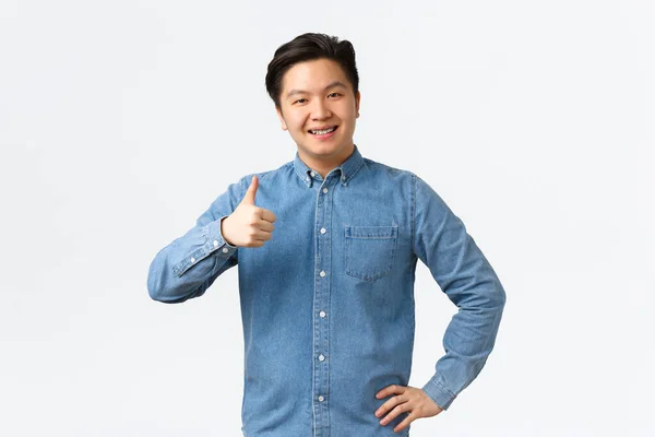 Usmívající se potěšený asijský muž student s rovnátka, ukazující palce nahoru, doporučit produkt nebo službu s vynikající kvalitou, jako a schválit nápad. Muž souhlasně přikyvuje, souhlasí s osobou, bílé pozadí — Stock fotografie