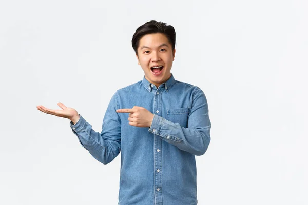 Sorrindo alegre bonito asiático homem fazendo anúncio, demonstrar novo produto na mão, apontando o dedo para espaço em branco para o seu anúncio, de pé divertido sobre fundo branco — Fotografia de Stock