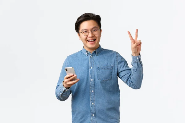 Χαρούμενος Ασιάτης με σιδεράκια, με γυαλιά και πουκάμισο, με κινητό τηλέφωνο, εφαρμογή smartphone, κάνει φιλειρηνική χειρονομία και χαμογελά στην κάμερα φιλικό, στέκεται λευκό φόντο — Φωτογραφία Αρχείου
