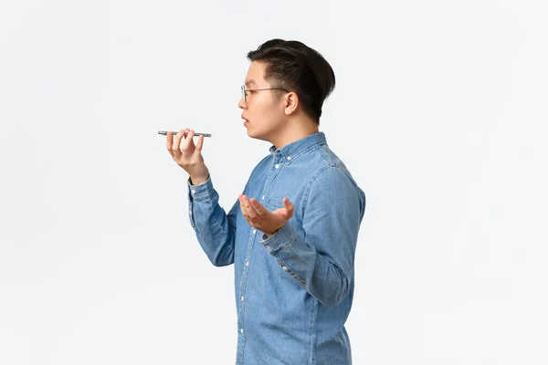 Profil av upptagen ung asiatisk manlig entreprenör, frilansare spela in röstmeddelande, prata i telefon högtalare, göra anteckningar på inspelare, ha samtal, stående vit bakgrund — Stockfoto