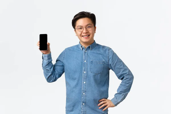 Sonriendo satisfecho asiático freelancer masculino, empresario con su propia pequeña empresa mostrando pantalla de teléfono inteligente complacido. Tipo con tirantes y gafas usando aplicación móvil, fondo blanco — Foto de Stock