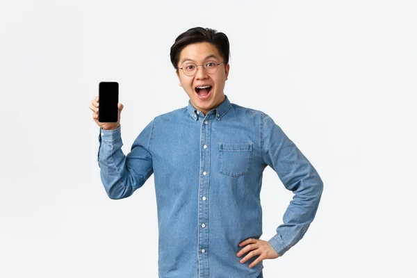 Glad och road stilig asiatisk kille med hängslen och glasögon reagerar på fantastiska nyheter, visar mobiltelefon skärm, införa ansökan eller butik, stående vit bakgrund förvånad — Stockfoto