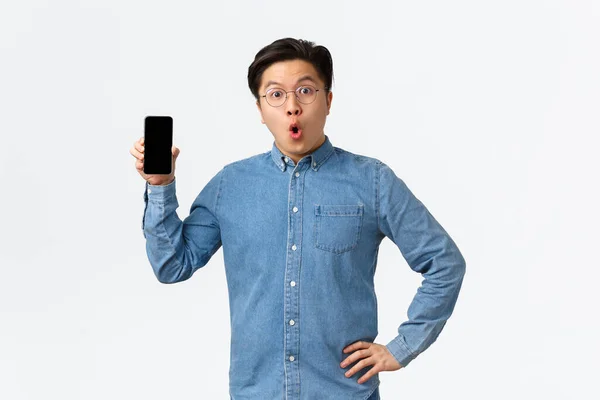 Surpreso e surpreso asiático homem em óculos e roupas casuais, mostrando tela do telefone celular e dizendo wow, discutindo novo aplicativo smartphone, última notícia on-line, fundo branco — Fotografia de Stock