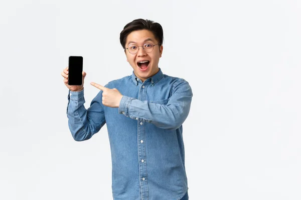 Spännande och imponerad asiatisk kille i glasögon och hängslen, visar awesome sak på mobilen skärmen, peka finger åt smartphone display som införa nya program, vit bakgrund — Stockfoto