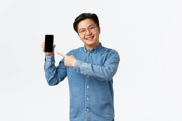Sorrindo bonito asiático cara com aparelho e óculos, apontando o dedo para tela do smartphone. Homem mostrando promo ou aplicativo na tela do telefone móvel, promover o site, de pé fundo branco — Fotografia de Stock