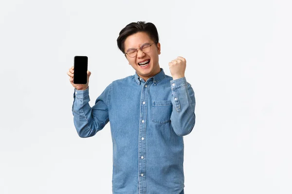 Vencedor bem sucedido asiático homem em óculos e aparelho, mostrando tela do smartphone e punho bomba em triunfo, show-off com pontuação no jogo móvel ou aplicação, de pé fundo branco — Fotografia de Stock