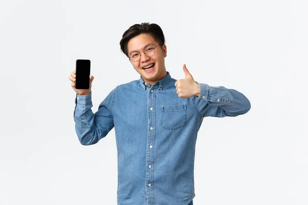 Upbeat e satisfeito asiático homem com aparelho, vestindo óculos e roupas casuais, mostrando thumb-up na aprovação do aplicativo smartphone, enfrentando tela do telefone móvel para promover aplicativo, fundo branco — Fotografia de Stock