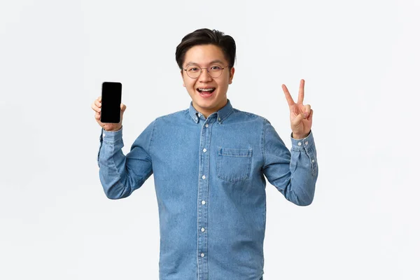 Sonriente alegre asiático freelancer masculino, estudiante en gafas y frenos, mostrando la pantalla del teléfono inteligente y signo de paz, de pie satisfecho con nuevos aplausos. Hombre recomienda descargar aplicación — Foto de Stock