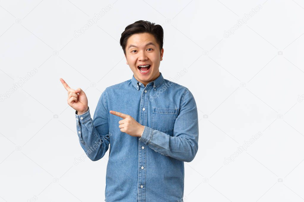 Excited cheerful asian man in blue shirt react to fantastic news, pointing fingers upper left corner, demonstrate banner, promote big event, smiling amazed, standing white background