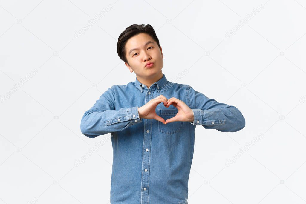 Silly handsome asian young guy in blue shirt confessing love, express sympathy, blogger grateful for followers, showing heart gesture and pouting as sending air kiss, white background