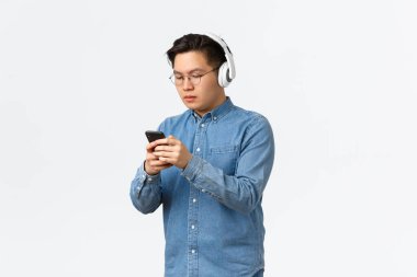 Yaşam tarzı, eğlence ve teknoloji konsepti. Ciddi görünümlü gözlüklü Asyalı genç adam, kablosuz kulaklıkla müzik dinliyor ve mesajlaşan arkadaşı, cep telefonu ayarlı kulaklık sesi kullanıyor.