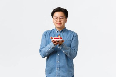 Kutlama, tatiller ve yaşam tarzı konsepti. Gözlüklü yakışıklı Asyalı adam dilek tutuyor ve doğum günü pastasına mum üflüyor, doğum gününü kutluyor, beyaz arka planda duruyor.