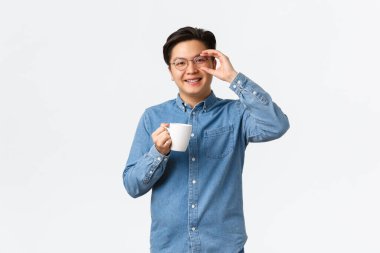 Gülümseyen Asyalı adam, bardaklı ofis çalışanı öğle yemeği molası veriyor, beyaz arka planda bir fincan kahveyle ayakta duruyor. Kupadan içen ve neşeli görünen bir adam, sabah rutini.