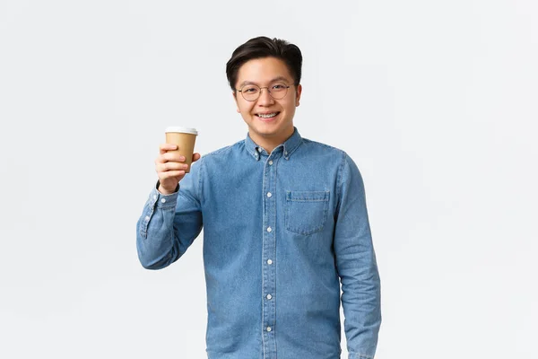 Livsstil, fritid och människor koncept. Le asiatisk manlig student i glasögon och hängslen höja kopp kaffe, gör jubel gest, dricka cappuccino takeaway från favorit café, vit bakgrund — Stockfoto