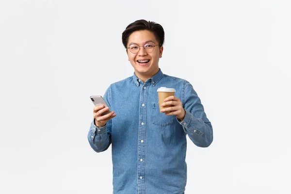 Estilo de vida, ocio y tecnología. Despreocupado hombre asiático sonriente con aparatos ortopédicos, beber café durante el almuerzo fuera de la oficina, el uso de teléfono móvil, amigos mensajes de texto, el envío de mensajes, fondo blanco — Foto de Stock