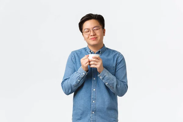 Avslappnad och glad leende asiatisk man med slutna ögon njuter av läcker doft av kaffe, hålla mugg och dricka cappuccino, morgon rutin och livsstil koncept — Stockfoto