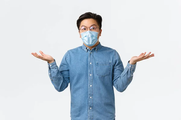 Covid-19, förebyggande av virus och social distansering på arbetsplatsen. Förvirrad och förvånad asiatisk manlig entreprenör sprida händerna i sidled och rycka förbryllad, bära medicinsk mask och glasögon — Stockfoto