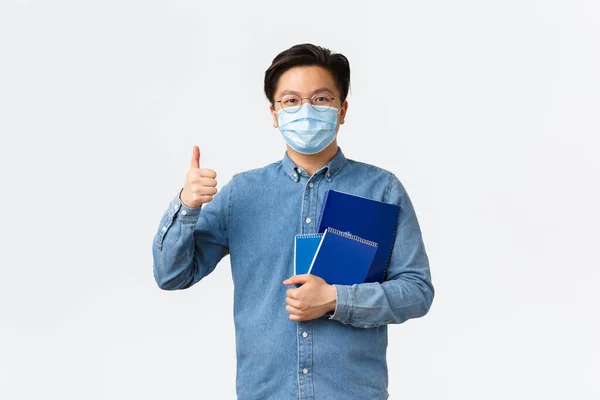 Covid-19, πρόληψη του ιού, και κοινωνική αποστασιοποίηση στο πανεπιστήμιο έννοια. Χαρούμενος ασιάτης δάσκαλος ή δάσκαλος με ιατρική μάσκα κουβαλάει σημειωματάρια και υλικά μελέτης, δείχνοντας αντίχειρες, λευκό φόντο — Φωτογραφία Αρχείου