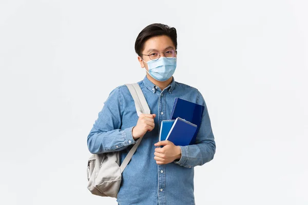 Covid-19, förebyggande av virus och social distansering på universitetskonceptet. Unga asiatiska manliga student i medicinsk mask bära ryggsäck och anteckningsböcker, förberedd för lektion, stående vit bakgrund — Stockfoto