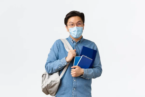 Covid-19, πρόληψη του ιού, και κοινωνική αποστασιοποίηση στο πανεπιστήμιο έννοια. Ξέγνοιαστος απερχόμενος ασιάτης φοιτητής, τύπος με ιατρική μάσκα και γυαλιά συζητώντας, κρατώντας σακίδιο και σημειωματάρια — Φωτογραφία Αρχείου