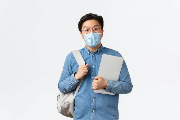 Covid-19, πρόληψη του ιού, και κοινωνική αποστασιοποίηση στο πανεπιστήμιο έννοια. Έκπληκτος απερχόμενος Ασιάτης, μαθητής με σακίδιο και φορητό υπολογιστή που ενδιαφέρεται για την κάμερα, φοράει ιατρική μάσκα και γυαλιά — Φωτογραφία Αρχείου