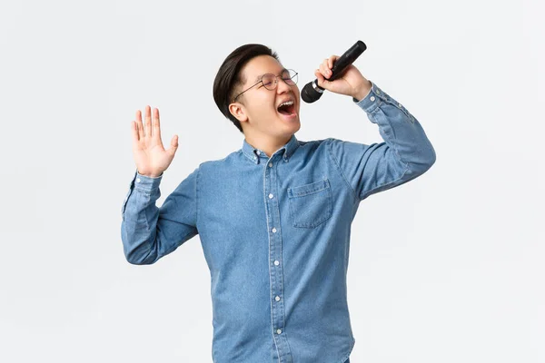 Lifestyle, vrije tijd en mensen concept. zorgeloze aziatische man in bril en beugel zingen lied in karaoke bar, holding microfoon, het uitvoeren van nieuwe single, staande witte achtergrond vreugdevol — Stockfoto