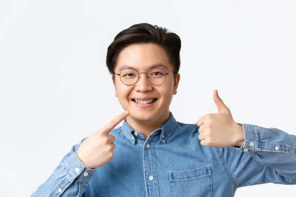 Ortodonti och stomatologi koncept. Närbild av nöjd asiatisk kille, tandvårdsklinik klient ler glad och pekar på hans tandställning och visar tummen upp i godkännande, rekommenderar — Stockfoto