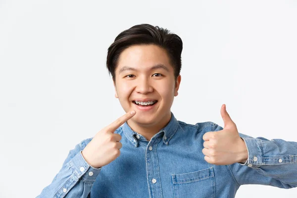 Orthodontie en stomatologie concept. Close-up van tevreden aziatische man, tandheelkundige kliniek klant glimlachend gelukkig en wijzend naar zijn gebitsbeugel en het tonen van duimen-up in goedkeuring, raden — Stockfoto